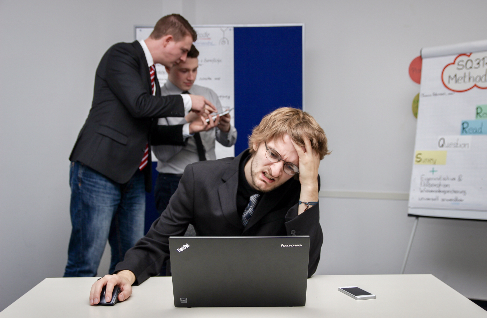 Stock image. The foreground has a man in the centre. He's working at a laptop looking stressed, left hand in hair and a grimace on his face. In the background behind the laptop man to our left are two gentlemen in smart casual dress looking at a calculator or something. Far right of theimage is a screenshot with generic token businessy words